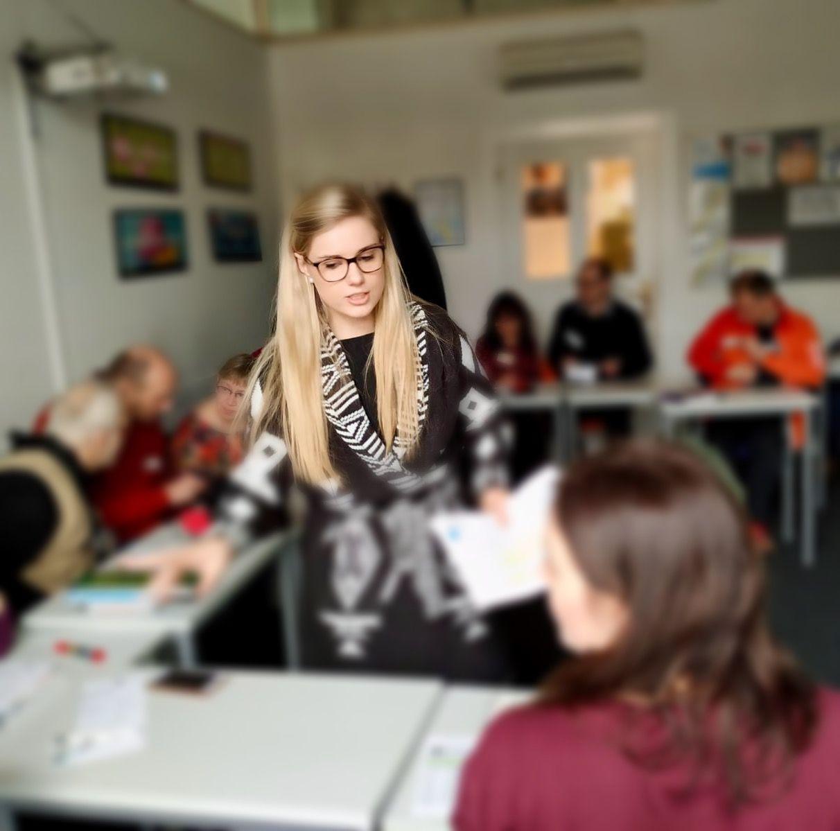 Woman with glasses leading a workshop in a classroom setting, engaging with attendees seated at tables.